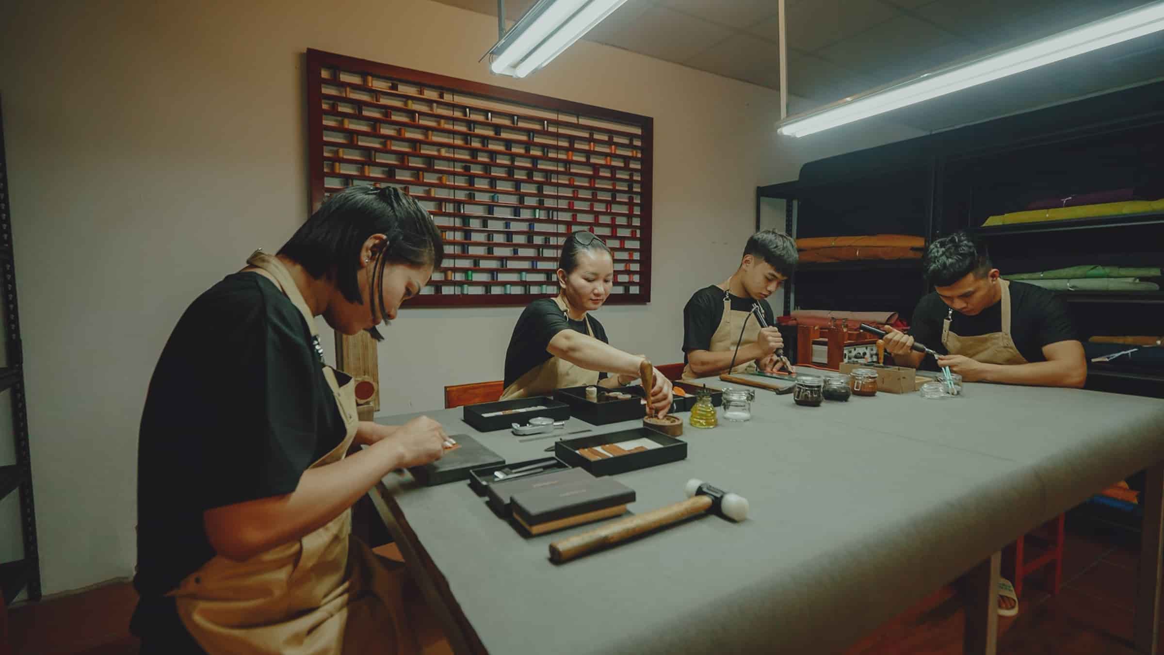 The AKC workshop in Vietnam: craftsmen at the long bench making leather watch straps, thread stock on the wall behind