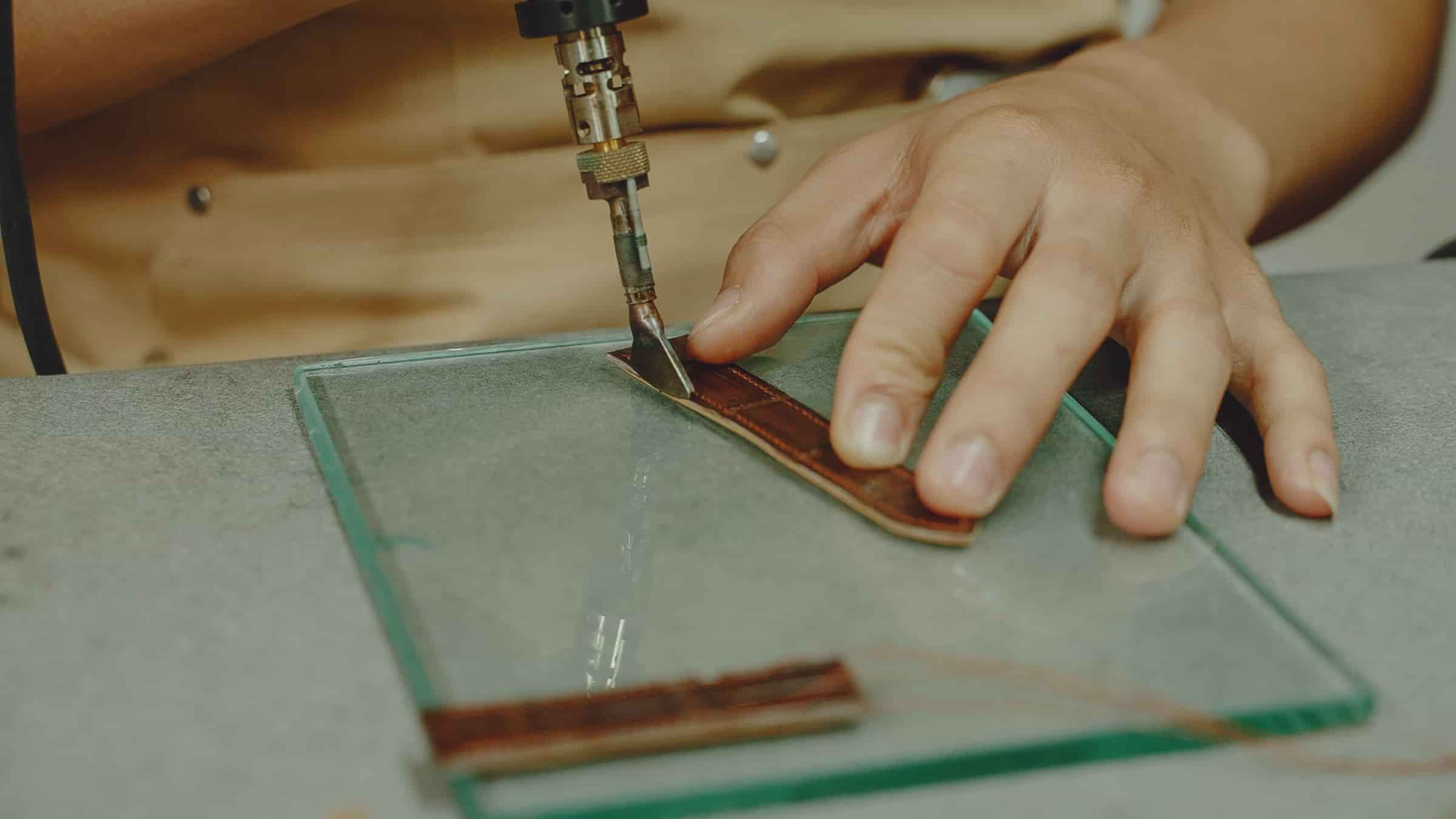 An AKC craftsman drawing a blade along a steel rule to cut a leather watch strap on a glass cutting plate