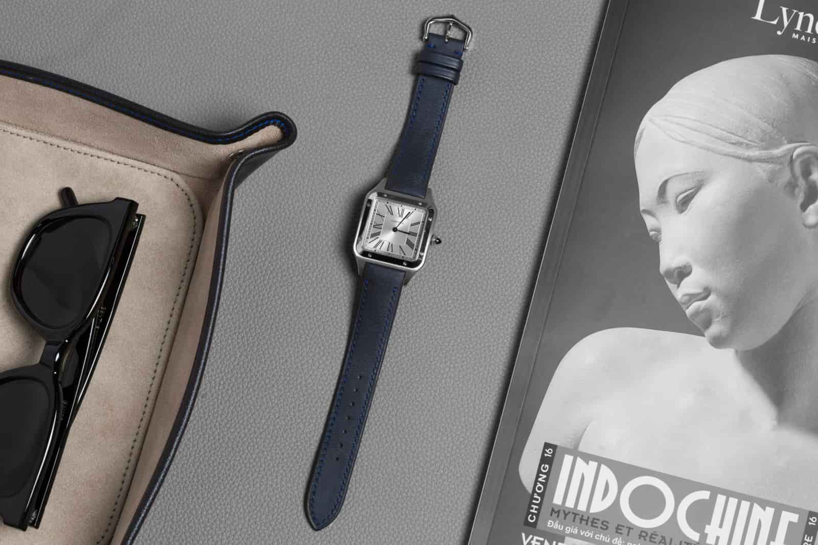Navy leather watch strap on a square-cased watch laid flat on a desk beside folded sunglasses and an open black-and-white magazine