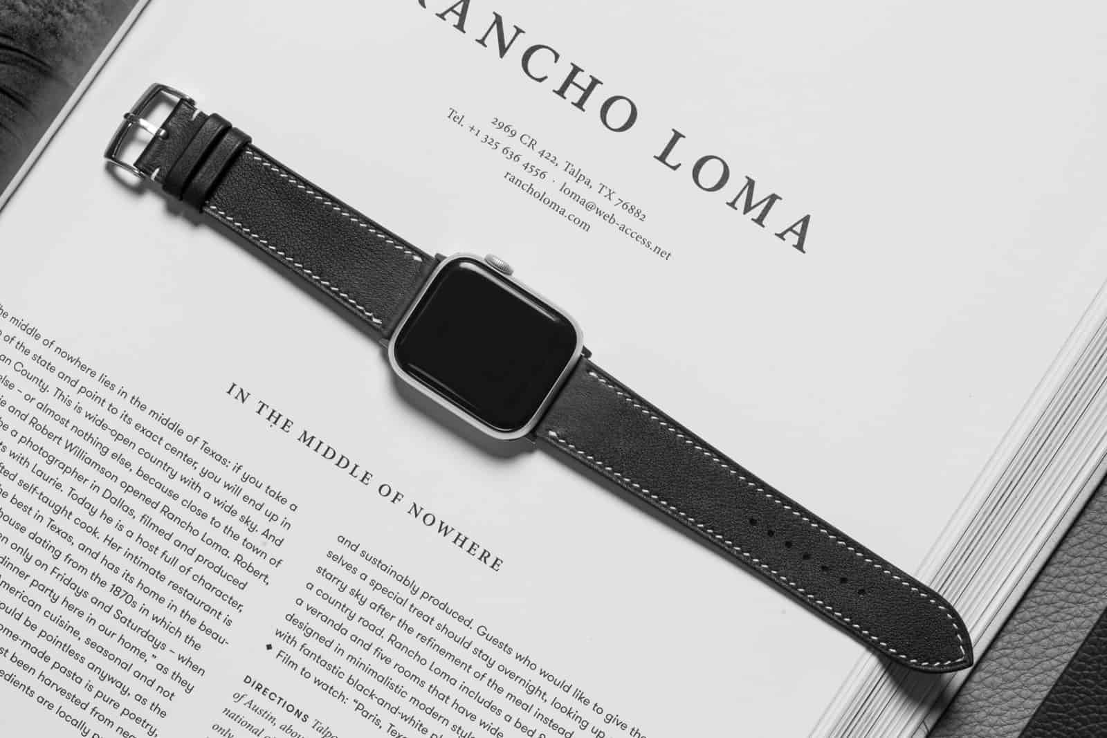 Black leather Apple Watch band lying across an open book, the full band, both adapters and the stitch line visible end to end